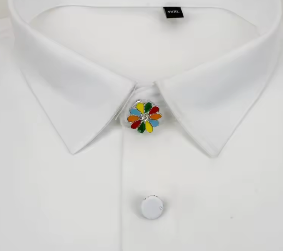 Färgglatt knappsmycke i blomform med emaljblad i rött, grönt, blått och gult samt klar sten i mitten, fäst på vit skjorta.
