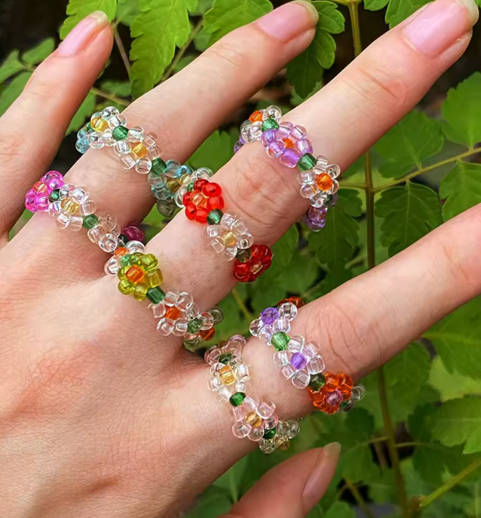Elastiska pärlringar med små blommor i flera färger, burna på fingrar mot grön bakgrund.
